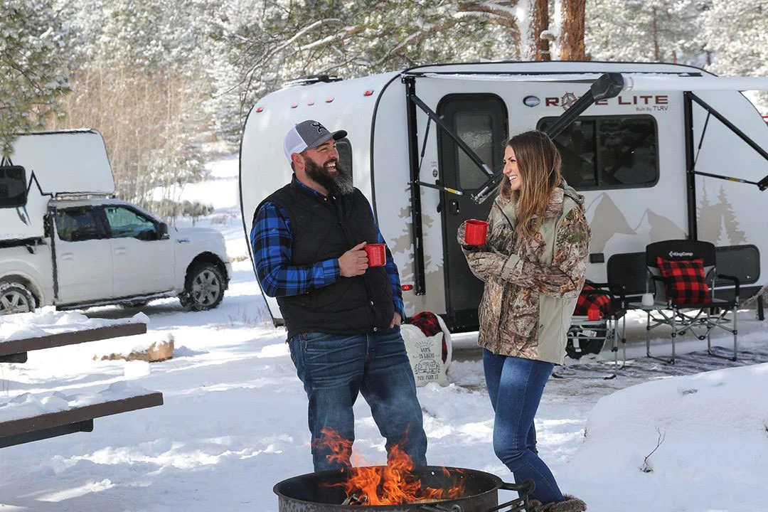 Two people around a fire, in the snow, with a camper in the background