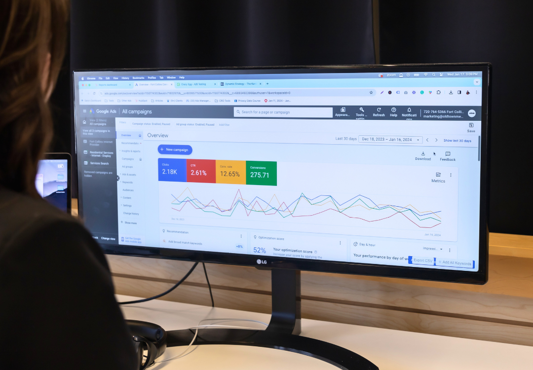 A person views a computer monitor displaying a Google Ads dashboard. The screen shows performance metrics and graphs for various campaigns, including clicks, impressions, and cost per click. The interface features navigation tabs and data visualization.