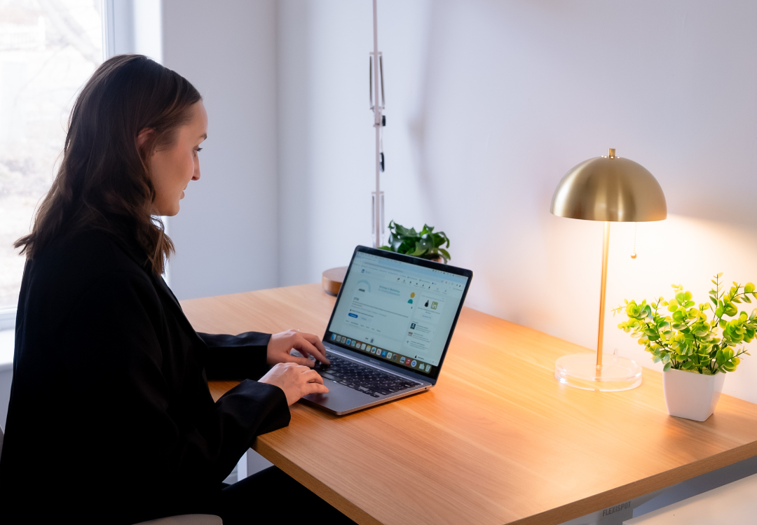 A person sitting at a wooden desk, working on a laptop. The desk features a gold lamp and small potted plants. Natural light is coming through a nearby window, illuminating the space. The person appears focused on the screen.