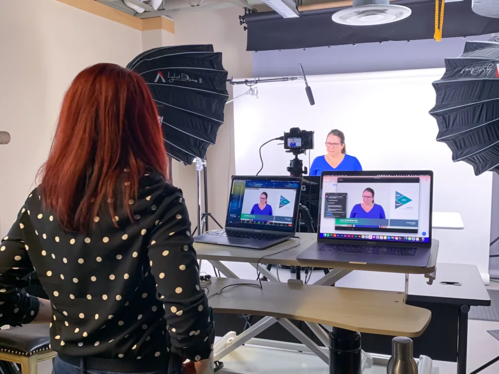 A woman with red hair stands at a desk looking at two laptop screens showing a woman in a blue top being recorded in a studio setup. The studio features professional lighting and a white backdrop.