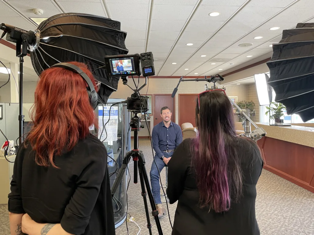 A man sits in an office under studio lighting, with two women filming him. The women, one with red hair and the other with long, dark hair with purple highlights, operate cameras and sound equipment. The setting appears professional with diffused lights and camera rigs.