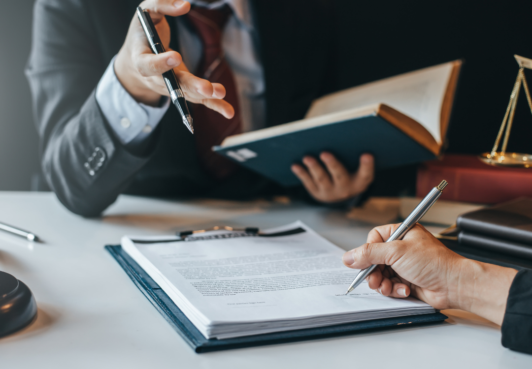 Two people are engaged in a discussion or meeting. One is holding an open book and gesturing with a pen, while the other is signing a document on a clipboard. A gavel and scales of justice are visible on the desk, indicating a legal or professional setting.