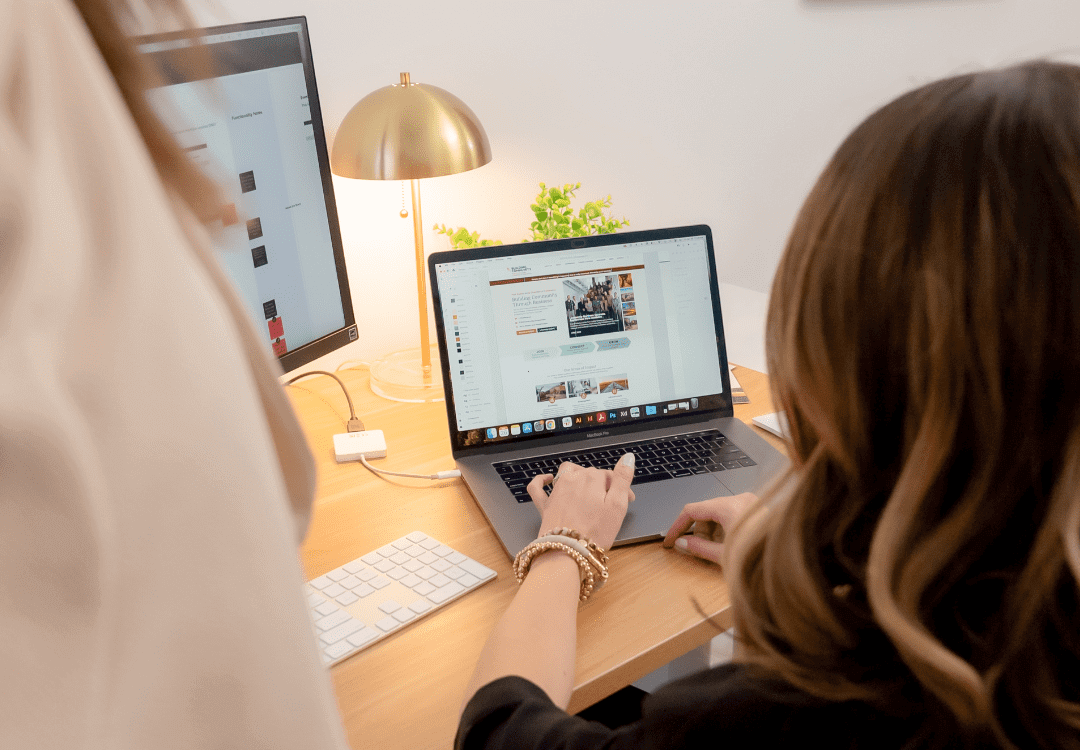 A woman with wavy hair, wearing a black top and gold bracelets, uses a laptop on a wooden desk. The screen displays a website. Another person, partially visible, stands nearby. The workspace includes a desk lamp, a small plant, and a secondary monitor.