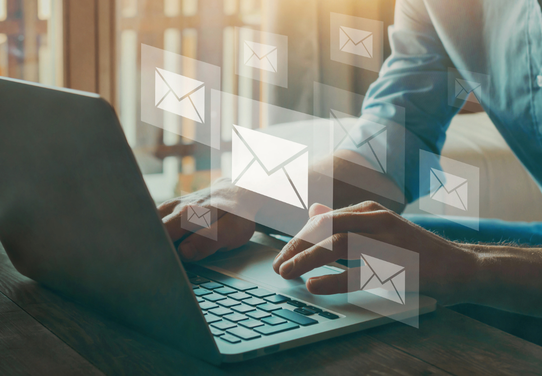 A person typing on a laptop with several email icons floating above the keyboard, symbolizing sending or receiving emails. The background is softly lit with natural light coming through a window, giving a warm ambiance.