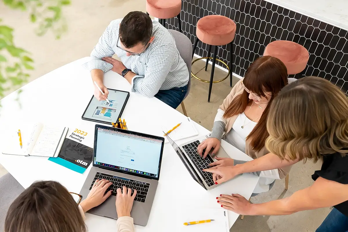 Four people are seated around a white table, collaborating on laptops and a tablet. They appear to be reviewing documents and discussing a project. The table has notebooks, pens, and a planner, while modern chairs and a bar counter with stools can be seen in the background.
