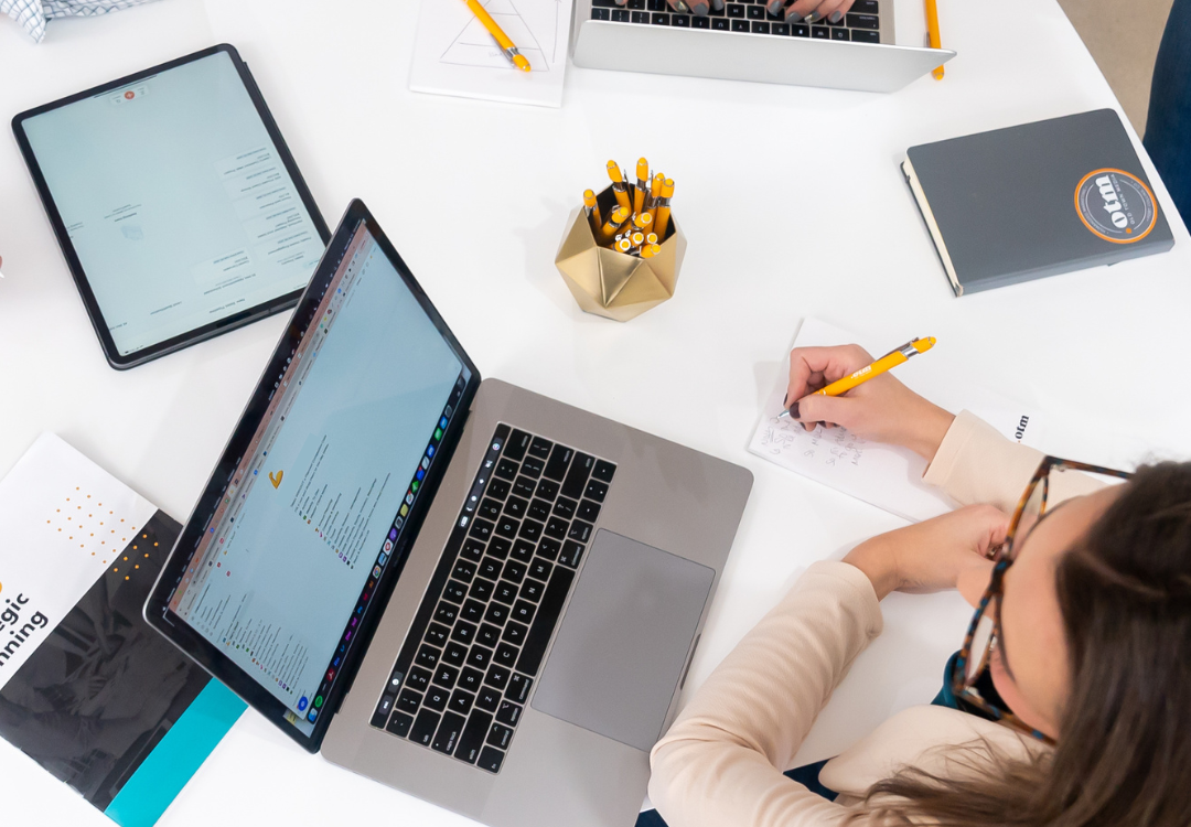 Top-down view of a workspace with a person writing on a notepad, surrounded by laptops, a tablet, a notebook, and a container of pencils. The person appears to be engaged in work or study, with documents and an open webpage visible on the devices.