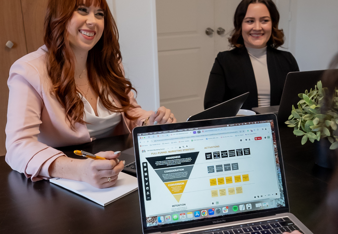 Two women are sitting at a table in an office setting, both with open laptops. One woman is writing in a notebook with a pen. In the foreground, there is a close-up of a laptop screen displaying a marketing strategy funnel diagram. A small plant is also on the table.