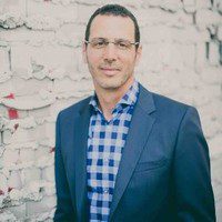 A man wearing glasses, a blue checked shirt, and a dark blazer stands in front of a white brick wall with red markings—looking at the camera and smiling slightly, he embodies the confidence of a scalable marketing engine for consulting firms.