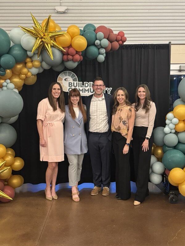 group-chamber Five people stand smiling in front of a “Building Community” sign, framed by an arch of colorful balloons and gold star decorations at an indoor event. Four women and one man are dressed in business or smart casual attire.