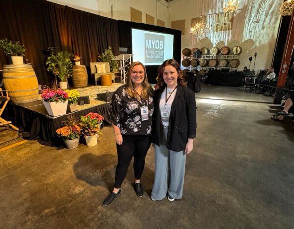 group-myob Two women wearing conference badges smile and stand together in a large event space. Behind them is a stage with plants, flowers, and a screen displaying "MYOB Mind Your Own Business.