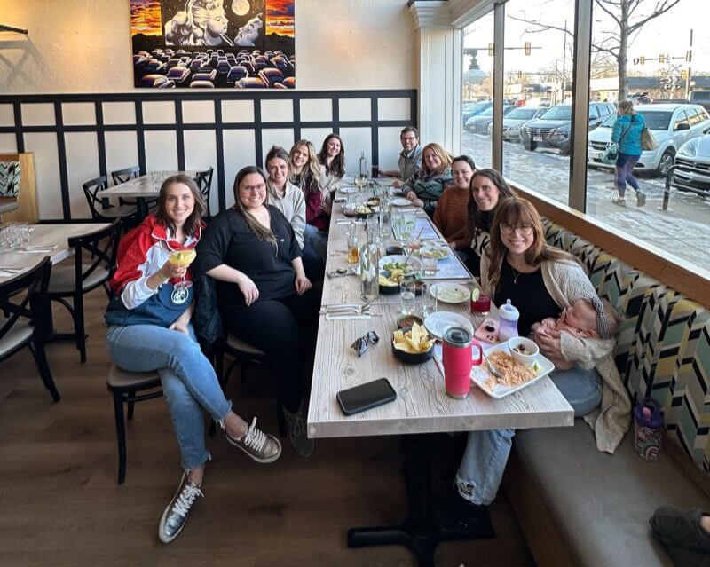 group-rio A group of ten people are sitting around a long restaurant table with food and drinks, smiling at the camera. One person holds a baby. Large windows show a street and cars outside. Artwork decorates the wall behind them.