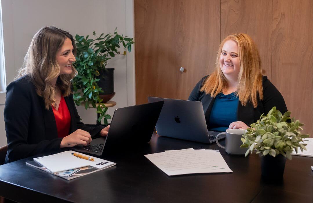 Two women sit at a table with laptops, notebooks, pens, and paperwork, smiling and talking in a modern office setting with plants and a coffee mug on the table.