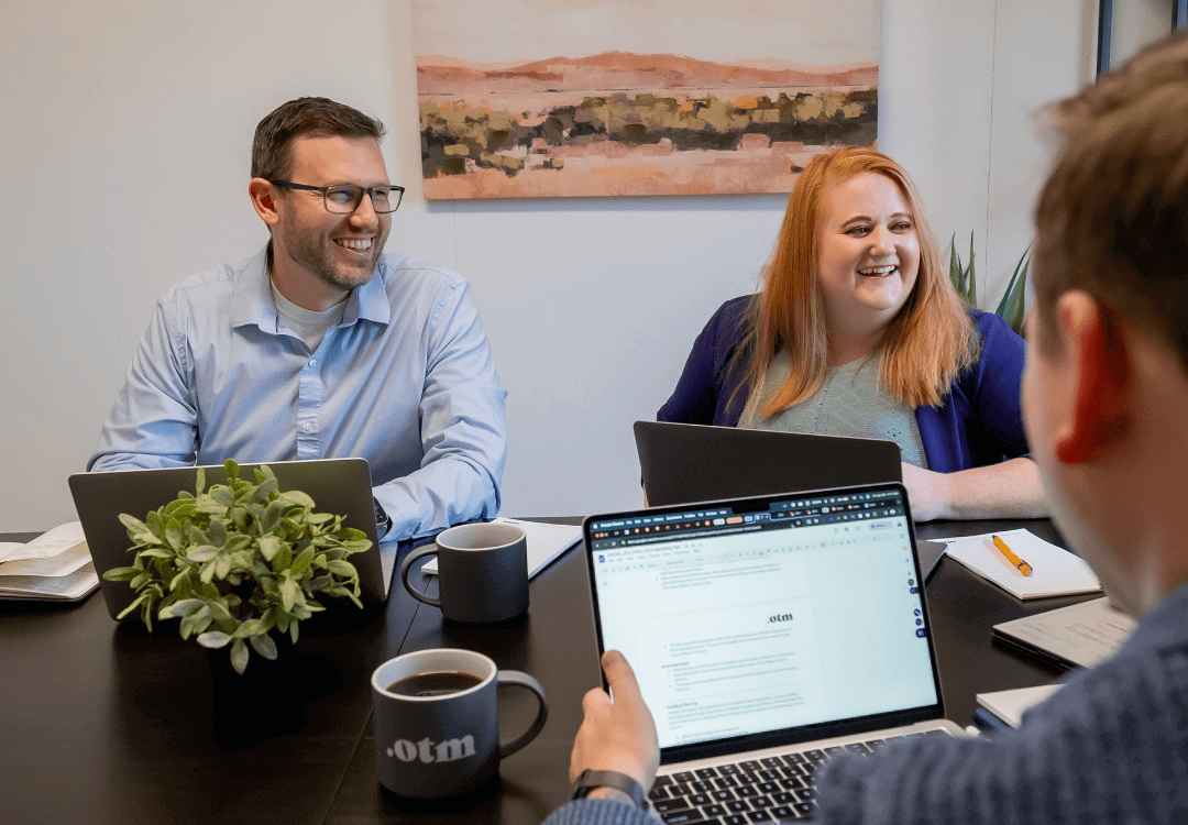 Three people sit at a table with laptops, smiling and talking during a meeting. A plant, coffee mugs, and notebooks are on the table. A landscape painting hangs on the wall in the background.