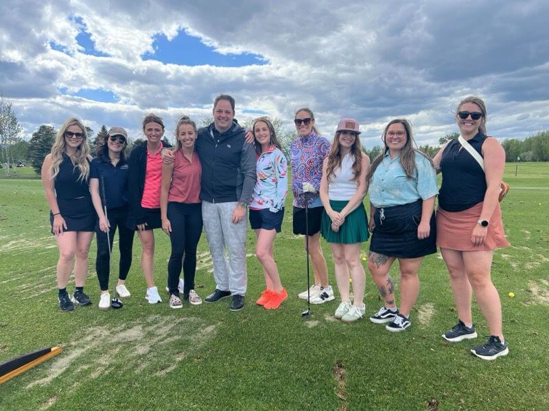 otm-team-golf A group of ten people stand together and smile on a grassy golf course under a cloudy sky. Some hold golf clubs, and they are dressed in casual and sporty outfits. Trees and greenery are visible in the background.