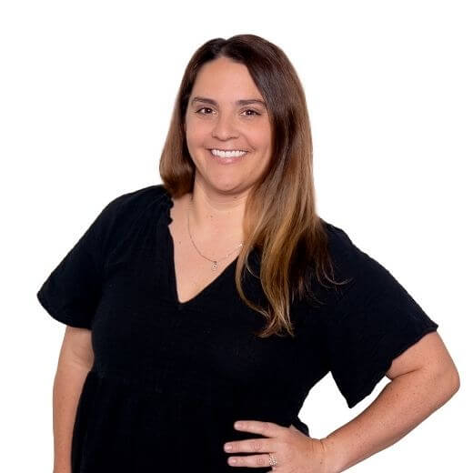 A woman with long brown hair wearing a black short-sleeve top stands smiling with one hand on her hip against a plain white background.