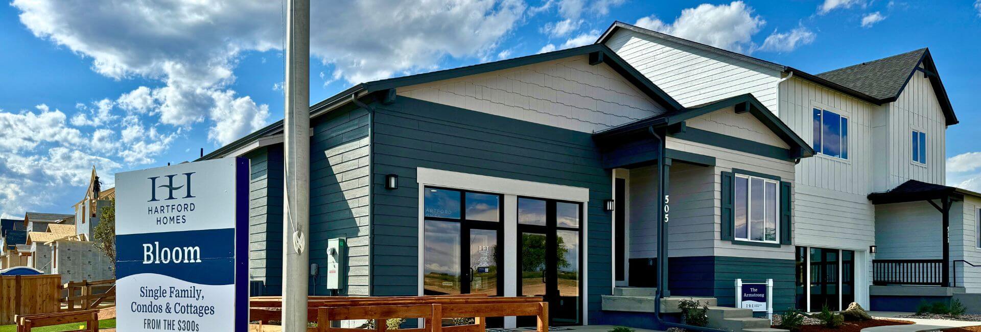 Modern two-story home with dark and light siding, large windows, and a wooden fence. A sign in front reads “Hartford Homes Bloom—Single Family, Condos & Cottages from the $400s.” Blue sky with clouds in the background.
