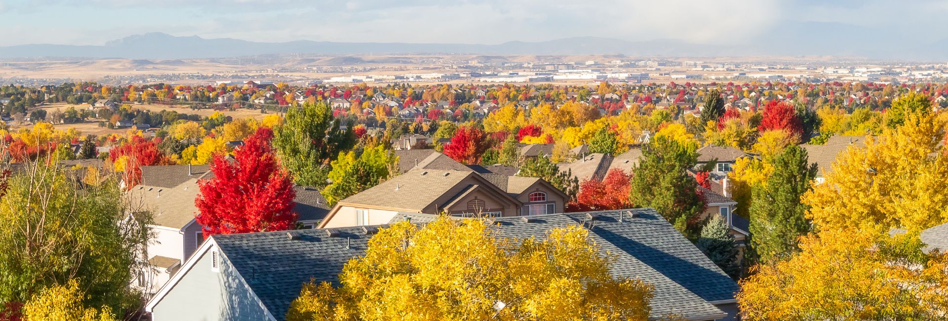 A suburban neighborhood in autumn, with houses surrounded by vibrant yellow, orange, and red trees; distant hills and a city skyline are visible in the background under a bright, partly cloudy sky.