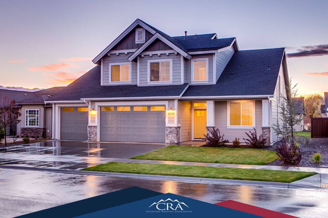 A modern two-story suburban house with gray siding, a three-car garage, and a well-kept lawn. The wet driveway reflects a sunset sky. The image supports a strong roofing industry content strategy with the Colorado Roofing Association logo at the bottom.