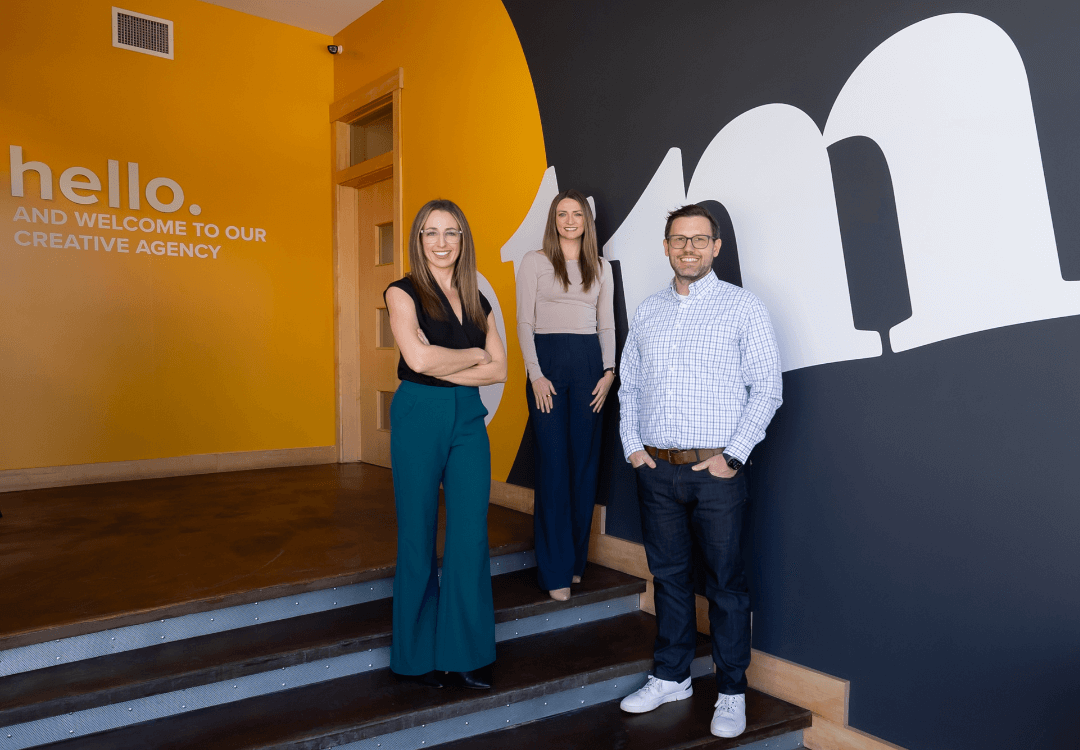 Three people stand and smile inside a modern office with yellow and black walls. A large "M" and the words "hello. and welcome to our creative agency" are displayed on the wall behind them.