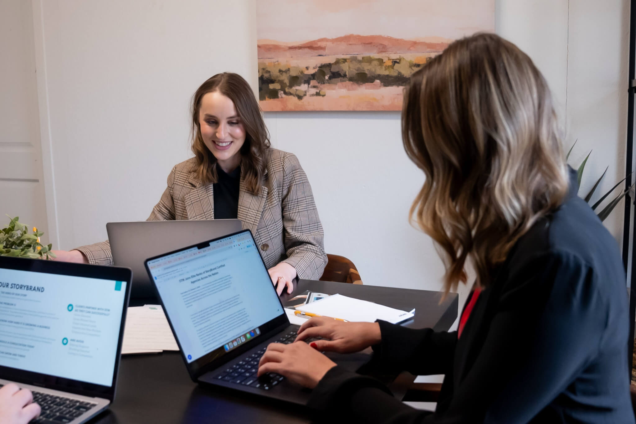 Two women sit at a table with laptops, working and smiling in a modern office setting. Documents related to sales and marketing alignment and a plant are on the table, with an abstract landscape painting hanging behind them.