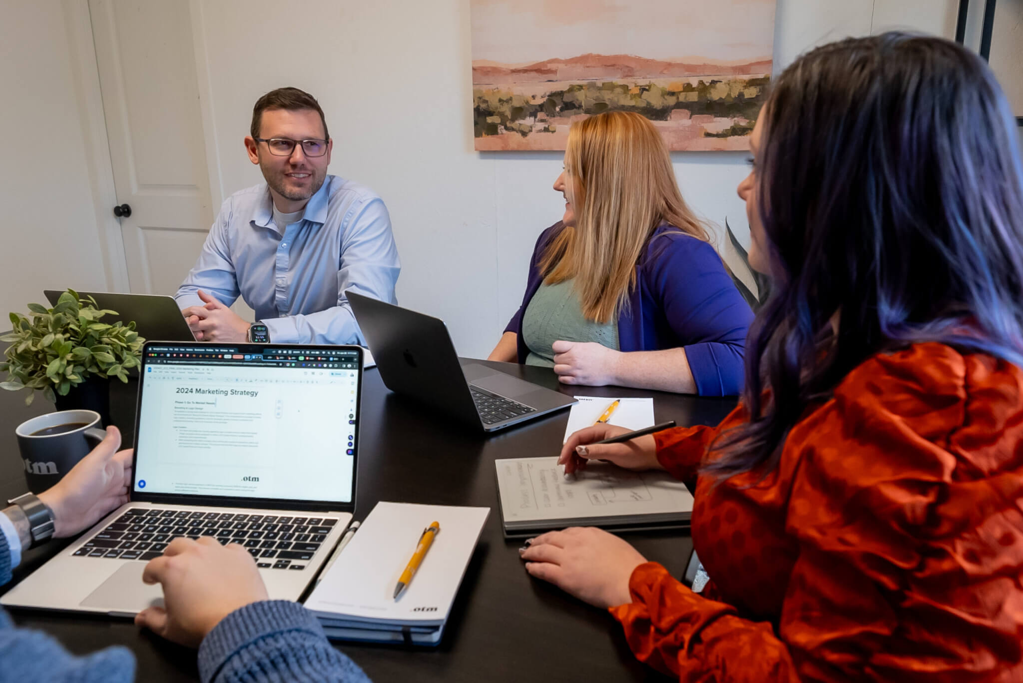 Four people sit around a conference table with laptops, notebooks, and coffee, engaged in a discussion. One person smiles while speaking; others listen and take notes. A landscape painting hangs on the wall behind them.