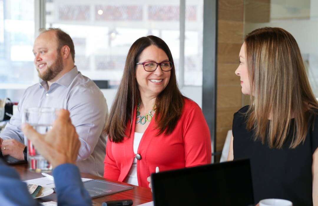 Three people sit at a conference table, smiling and engaged in conversation. The woman in the center wears glasses and a red cardigan. A laptop and drinks are visible on the table in front of them.