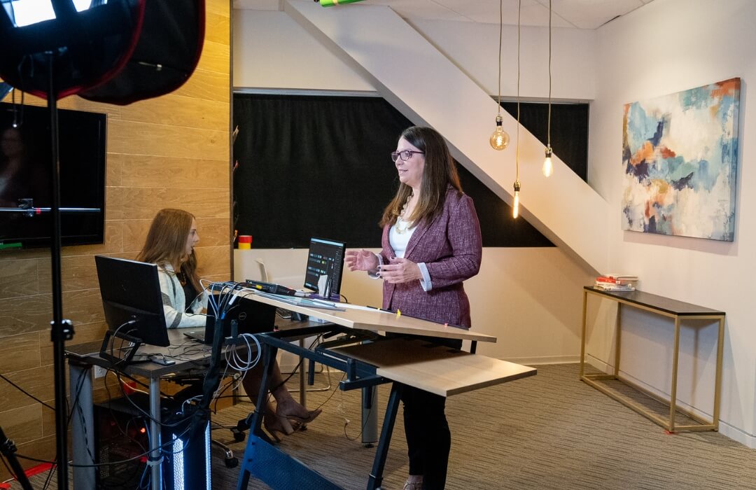 Two women work in a modern office studio with computers and lighting equipment. One stands and speaks while the other sits at a desk with technical gear. Abstract art and hanging lights decorate the space.