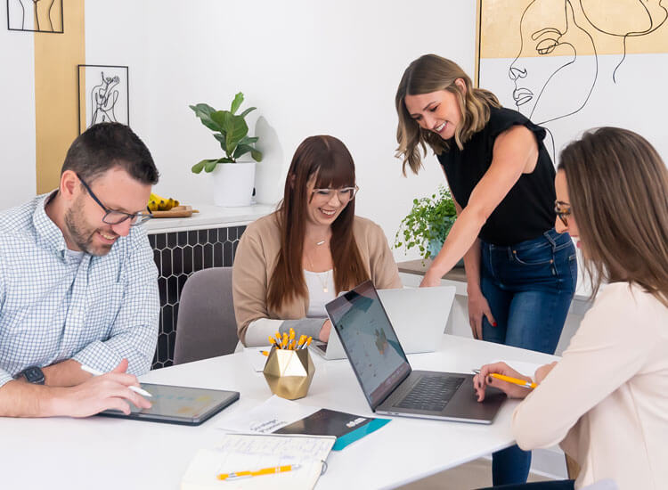Four people sit around a white table in a bright office, working on laptops and tablets. One woman stands, smiling and pointing at a computer screen, while others engage and take notes. Art and plants decorate the background.