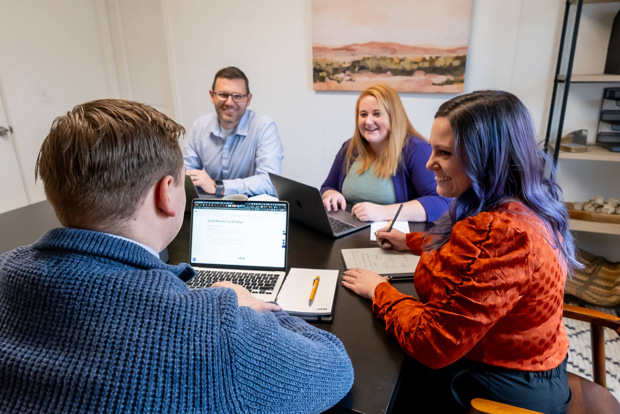 Four people sit around a table in an office, engaged in a meeting. Two men and two women are smiling, with laptops, notebooks, and pens in front of them. A landscape painting hangs on the wall behind them.
