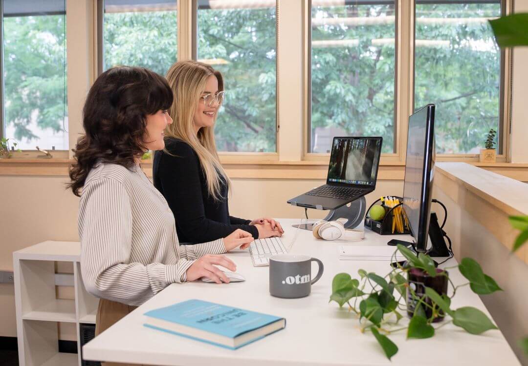 Two women stand at a desk in a bright office, collaborating on computers. Plants, a mug, and a book sit on the white desk. Large windows reveal greenery outside—a modern space reflecting AI adoption in professional services firms.