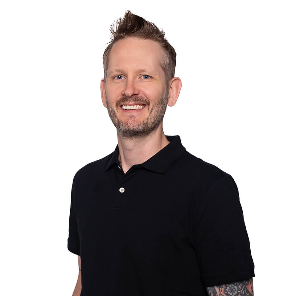 otm-ian-dougan Smiling man with short, light brown hair and a beard, wearing a black polo shirt. He has visible tattoos on his arm and is standing against a plain white background.