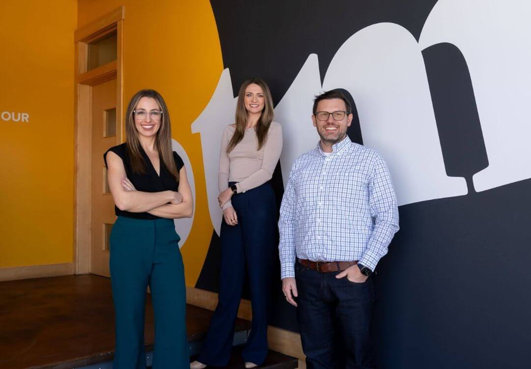 OTM leadership team members Vallene Kailburn, Kerrie Luginbill, and Miles Kailburn pose together inside the company office. Standing in front of a bold black, white, and yellow branded wall mural, the three smile confidently, representing the agency’s leadership team and professional services expertise.